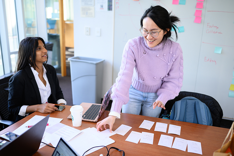  A woman in a white shirt and black blazer sits at a table, collaborating with a woman in jeans and a pink sweater who is standing at the table.