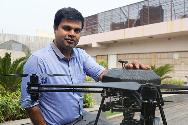  A man in a blue shirt poses with a black drone in a courtyard.