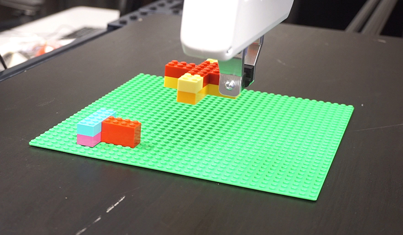  A white robot arm places an X-shaped figure made of Lego blocks on a large green Lego base.