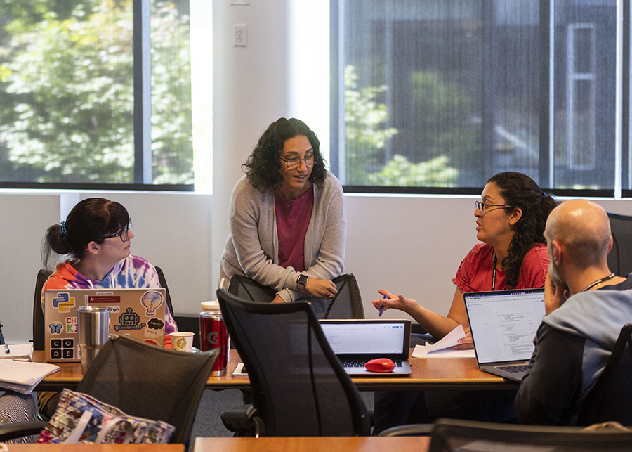  A woman leans her arms on a chair at a conference room table while women seated on her right and left look on. The back of a man's head is visible in the lower right corner.