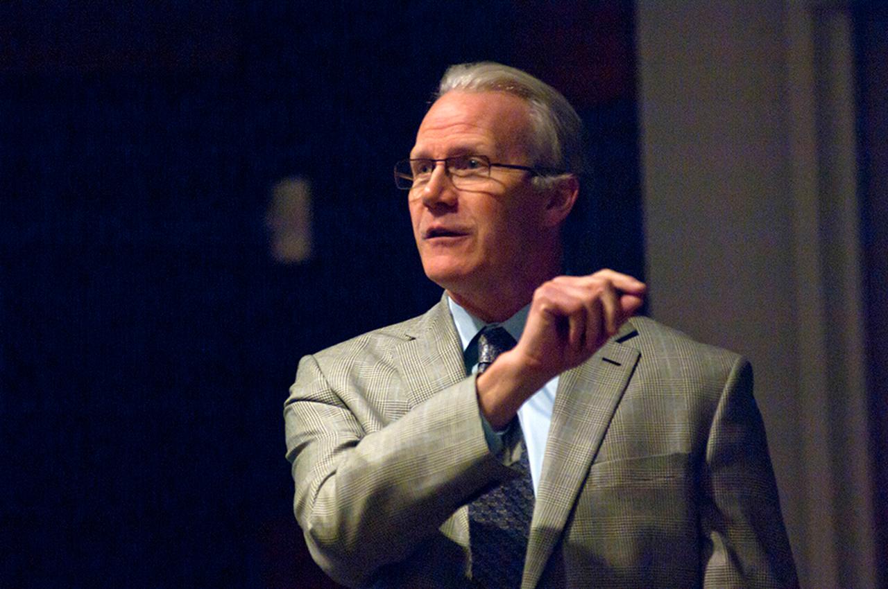  Randy Bryant speaks before an out-of-shot crowd. He has gray hair and wears a tan suit coat with a light blue line throughout and a blue shirt.