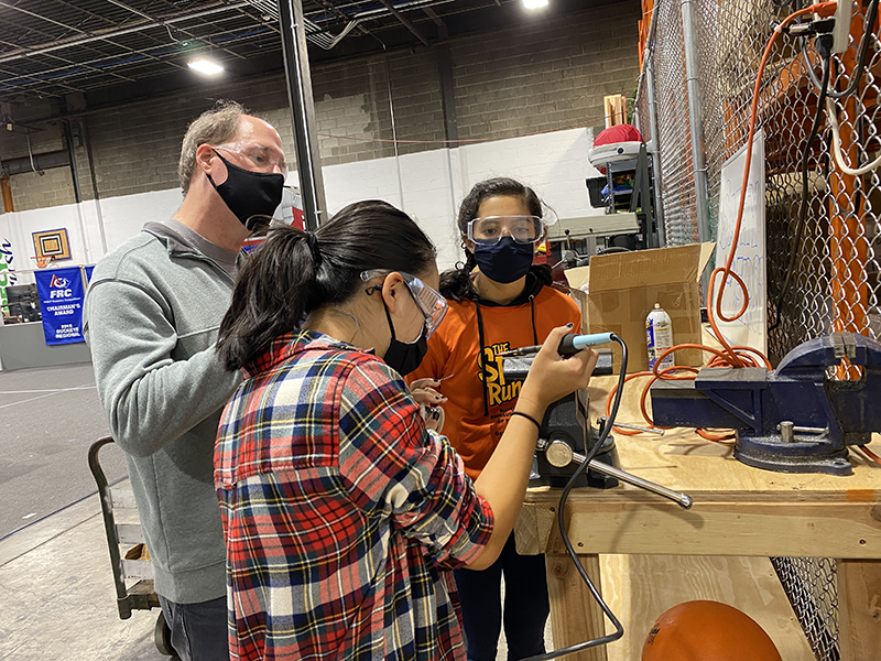  Two young girls, both with dark hair, use a soldering iron under the guidance of an adult man wearing a grey pullover with a facemask and safety glasses.