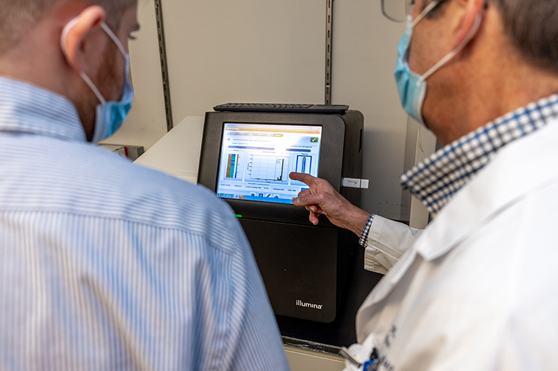  A gentleman in a striped dress shirt and mask looks at a computer monitor with another gentleman in a white coat and mask.