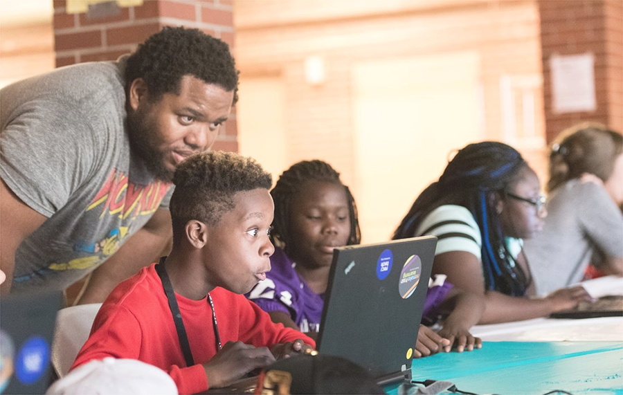  Students work on laptops in a classroom while an adult looks over their shoulders to help.