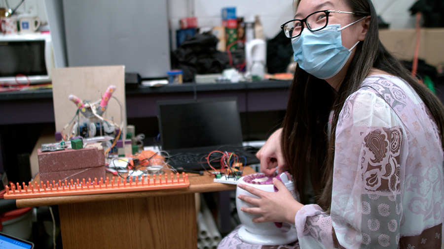  A woman in glasses wearing a pink and lavendar top holds a small white drum-shaped item with pink fabic woven on the top. Bricks, a laptop and knitting paraphernalia litter a table behind her.